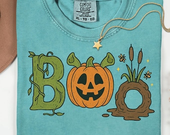 May include: A turquoise Comfort Colors t-shirt with a Halloween-themed graphic. The design features the word "BOO" with a green vine-like "B", an orange jack-o'-lantern, and a brown marsh scene. A gold necklace with a star pendant rests on the shirt.