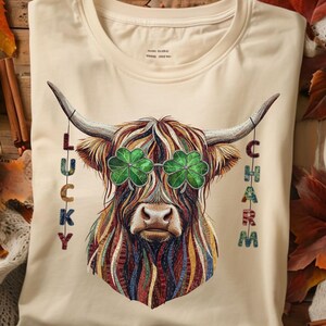 Könnte beinhalten: Cremefarbenes T-Shirt mit einer Highland-Kuh mit Kleeblatt-Brille und den Worten "LUCKY CHARM" vertikal auf beiden Seiten. Die Kuh hat mehrfarbiges Fell und große Hörner. Das Shirt liegt auf einer Holzoberfläche.