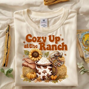 May include: Cream-colored t-shirt with the text "Cozy Up at the Ranch" in brown. The graphic includes a sunflower, hay bales, pumpkins, and a mug of coffee with whipped cream. A rustic, fall-themed design.