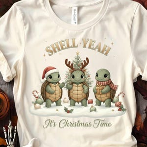 Puede incluir: Camiseta color crema con un gráfico festivo. El diseño presenta tres tortugas de dibujos animados con atuendo navideño, con el texto "SHELL YEAH" arriba y "It's Christmas Time" abajo. Una tortuga lleva un gorro de Papá Noel, otra tiene cuernos y la tercera una bufanda.