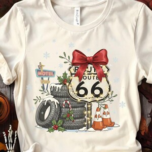 May include: Cream-colored t-shirt featuring a festive Route 66 design. The graphic includes a red bow, a Route 66 sign, tires, and a motel sign, all decorated with Christmas elements. The design also includes snowflakes and candy canes.