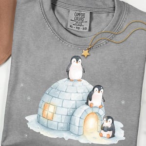 May include: A gray Comfort Colors t-shirt featuring a watercolor illustration of three penguins and an igloo. A gold-colored necklace with a star pendant is draped across the shirt. The shirt has a tag that reads XL - TG - EG.