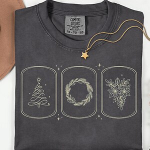 May include: Dark grey XL Comfort Colors t-shirt with three framed holiday-themed illustrations in cream. A Christmas tree, wreath, and floral design are featured. A gold star pendant necklace is draped across the shirt.