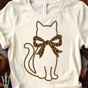 May include: Cream-coloured t-shirt featuring a cat outline made of brown beads. The cat has a leopard print bow. The shirt is made of a soft material and is suitable for casual wear.