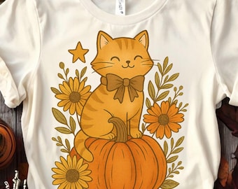 May include: Cream-colored t-shirt featuring a cartoon cat with orange and brown stripes, wearing a bow tie, sitting on an orange pumpkin. The design includes sunflowers, leaves, and a star, perfect for fall.