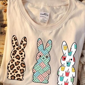 Può includere: T-shirt color crema con tre disegni di coniglietti. Un coniglietto ha una stampa leopardata, un altro un motivo pastello intrecciato e il terzo è ricoperto da illustrazioni di bevande colorate. La maglietta ha un'etichetta con la scritta "Comfort".