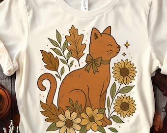 May include: Cream-colored t-shirt featuring a brown cat with a bow tie, surrounded by sunflowers and autumn leaves. The cat is smiling, and the design has a vintage, fall-themed aesthetic. The shirt is made of a soft material.