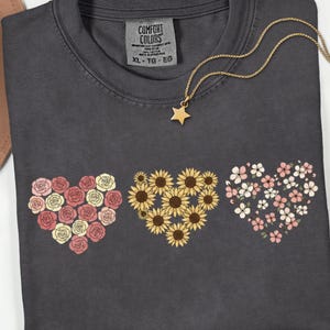 May include: Dark gray Comfort Colors t-shirt with three floral heart designs: roses, sunflowers, and small pink flowers. A gold star pendant necklace rests on the shirt. The tag reads XL-TG-EG.