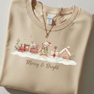 May include: Beige t-shirt with a Christmas-themed graphic. The design includes a red train, a teddy bear wearing a Santa hat and scarf, wrapped gifts, and a gingerbread house. The text "Merry & Bright" is printed below the graphic.