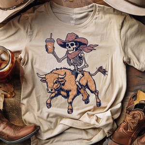 以下が含まれることがあります： ベージュ色のTシャツ。カウボーイの骸骨がコーヒーカップを持ち、雄牛に乗っているイラスト。骸骨はカウボーイハットと赤いバンダナを着用。雄牛は茶色で青色のアクセント。ヴィンテージ、ウエスタンテーマ。
