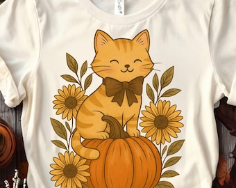 May include: Cream-colored t-shirt featuring a cartoon cat with a brown bow tie sitting on an orange pumpkin. The cat is surrounded by sunflowers and leaves, creating a fall-themed design.