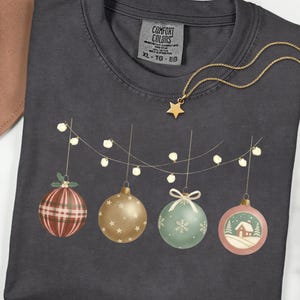 May include: Dark gray Comfort Colors XL t-shirt with a Christmas ornament design. The design features four ornaments in red plaid, gold stars, mint green snowflake, and a pink winter scene. A gold star necklace is draped on the shirt.