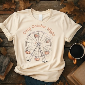 May include: Cream-colored t-shirt featuring a Ferris wheel design with pumpkins and ghosts. The text "Cozy October Night" is arched above the wheel. The shirt is displayed on a wooden surface with fall leaves and a coffee mug.