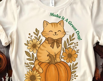 May include: Cream-colored t-shirt featuring a cartoon cat with a bow tie sitting on a pumpkin, surrounded by flowers and foliage. The text "Make it a Great Day!" is arched above the cat. The design has a fall theme.