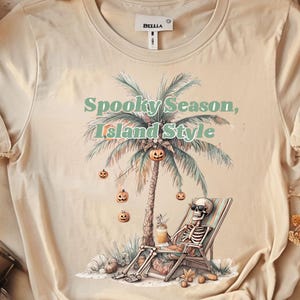May include: Beige t-shirt with a graphic of a skeleton relaxing on a beach chair under a palm tree with jack-o'-lanterns. The text "Spooky Season, Island Style" is above the palm tree. The shirt has a crew neck and short sleeves.