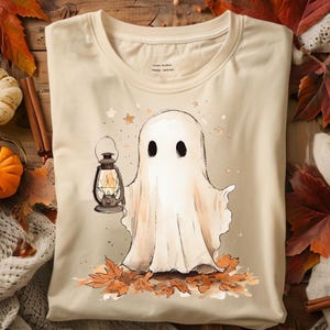 May include: Beige t-shirt featuring a watercolor illustration of a friendly ghost holding a lantern. The ghost stands on a bed of autumn leaves with stars in the background. A perfect tee for Halloween or fall.