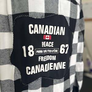 May include: Black and white plaid shirt with a large black patch. The patch features the word "CANADIAN" above a Canadian flag, with the words "PEACE", "18", "67", "FREEDOM", and "CANADIENNE".