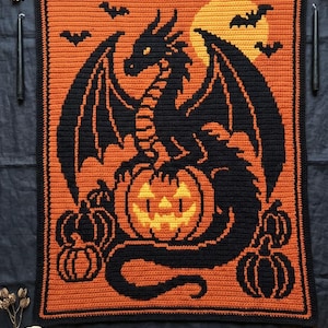 Puede incluir: Una manta de ganchillo rectangular que presenta un dragón negro posado sobre una gran calabaza de Halloween. El fondo es naranja con una luna llena amarilla y siluetas negras de murciélagos volando. Calabazas negras pequeñas en las esquinas.