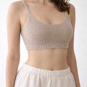 以下が含まれることがあります： ベージュのかぎ針編みクロップトップ、細いストラップ付き。明るい色のパンツと組み合わせています。かぎ針編みのステッチがテクスチャード加工の外観を作り出しています。全体的なスタイルはカジュアルで快適で、暖かい気候に適しています。