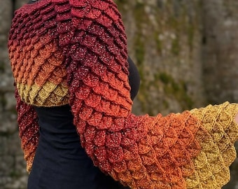 May include: A crochet shrug featuring a textured scale pattern in a gradient of colors transitioning from deep burgundy at the shoulders to bright orange and golden yellow at the flared sleeves, worn over a plain black short-sleeved shirt.