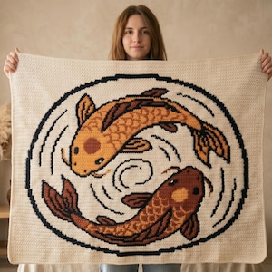 May include: A rectangular cream-coloured crochet blanket featuring a large circular design in the centre. The design depicts two koi fish in shades of orange and brown, swimming in a circular motion, enclosed by a thick black circular border.
