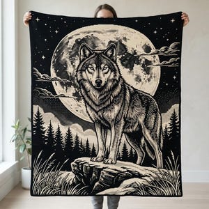 May include: A rectangular woven throw blanket featuring a high-contrast black and cream graphic design. The illustration depicts a wolf standing on a rocky outcrop in front of a large full moon, surrounded by a forest of pine trees and starry sky.