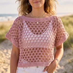 May include: A light pink crochet top with a honeycomb pattern. The top has short sleeves and a round neckline. A white tank top is visible underneath. The background shows a beach scene with the ocean and sand.