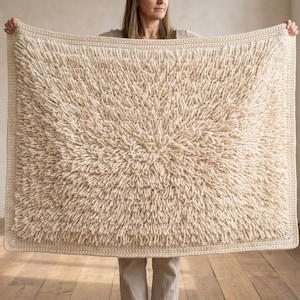 May include: A rectangular cream-colored crochet blanket featuring a dense, shaggy loop-stitch texture across the center and a flat, solid crochet border framing the entire piece. The yarn is a light off-white shade with a consistent, thick pile.