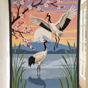 Op de afbeelding: Een rechthoekige gehaakte deken met een landschap van twee roodkruinkraanvogels, een berg, een grote witte zon en kersenbloesemtakken. Het ontwerp heeft een donkergrijze rand en is uitgevoerd in tinten blauw, roze en wit.