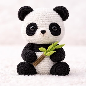 Puede incluir: Un oso panda de ganchillo hecho a mano, en blanco y negro. El panda sostiene una pequeña rama de bambú con hojas verdes. El panda tiene ojos, orejas y patas negras. El panda está sentado sobre una superficie blanca.