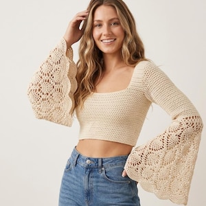 Op de afbeelding: Een beige gehaakte crop top met een vierkante halslijn en lange, uitlopende klokmouwen met een ingewikkeld, opengewerkt cirkelpatroon. Het lijfje is gemaakt met een dichte, vaste haaksteek, terwijl de mouwen overgaan in een decoratief kantontwerp met geschulpte randen.