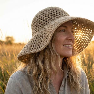 May include: A beige crochet sun hat with a wide brim. The hat is worn by a person with blonde hair, smiling in a field of tall grass. The sun shines through the hat's open weave, creating a warm, natural aesthetic.