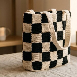 May include: A handmade tote bag with a black and cream checkerboard pattern. The bag is made of a thick, textured yarn and has two cream-colored handles. The bag is sitting on a neutral-colored surface.