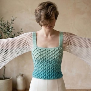 Puede incluir: Un top sin mangas con cuello cuadrado y un patrón de color degradado, que va del verde al azul. El top presenta mangas anchas de tejido abierto. La prenda se combina con pantalones blancos.