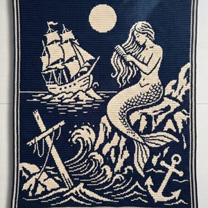 May include: A rectangular crochet blanket featuring a dark navy blue background with cream-colored yarn detailing. The design depicts a mermaid sitting on a rock, a sailing ship on the water, a full moon, crashing waves, and a ship anchor.