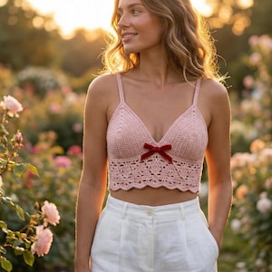 May include: A light pink crochet crop top with a red velvet bow. The top features a delicate, lacy design and thin straps. The model is wearing white trousers. The background is a rose garden.