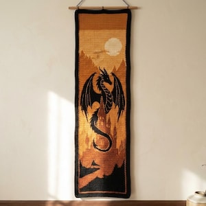 May include: A rectangular crochet wall hanging featuring a black dragon silhouette centered over a castle. The tapestry uses shades of orange, yellow, and brown to depict a sunset landscape with a full moon. It is suspended from a wooden rod.