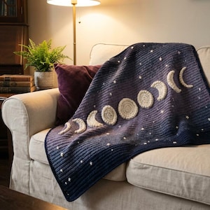Può includere: Una coperta all'uncinetto con uno sfondo sfumato dal blu scuro al viola, caratterizzata da una serie di fasi lunari color crema e piccole stelle bianche ricamate, con un motivo a coste testurizzato su tutta la superficie.