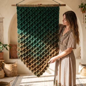 May include: A large, rectangular crochet wall hanging featuring a textured scale pattern. The yarn transitions in color from a deep forest green at the top to a warm, earthy brown at the bottom. It is suspended from a horizontal wooden dowel.