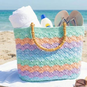 Pode incluir: Uma colorida bolsa de praia de crochê com alça de bambu, preenchida com uma toalha branca enrolada, protetor solar e chinelos. A bolsa apresenta listras em tons pastel de azul, pêssego e lavanda. A cena se passa em uma praia arenosa.