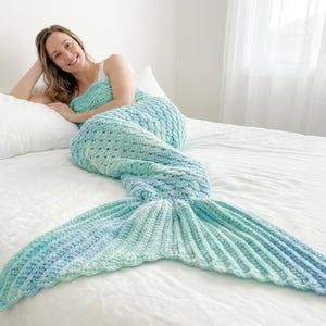 Peut inclure: Une couverture en crochet en forme de queue de sirène avec un motif de couleurs variées en bleu clair, turquoise et blanc. La couverture présente un point texturé en forme d'écailles sur le corps et une nageoire côtelée en éventail.