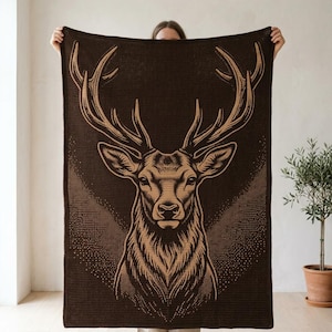 May include: A rectangular dark brown knit blanket featuring a large, detailed tan graphic of a stag's head with prominent antlers. The design is centered on the fabric, showcasing intricate line work that defines the animal's facial features.