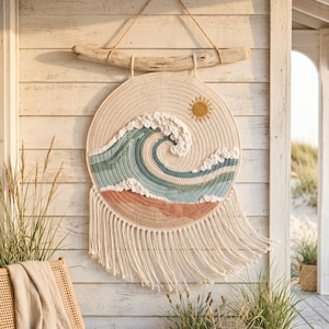 May include: A circular crochet wall hanging featuring a stylized ocean wave design in shades of cream, light blue, and terracotta. The piece includes a textured sun, a driftwood hanger with a rope cord, and long cream-colored fringe at the bottom.