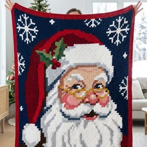 May include: A rectangular crochet blanket featuring a large portrait of Santa Claus wearing a red hat with white trim and holly leaves. The background is dark blue with white snowflake patterns, and the blanket has a solid red border.