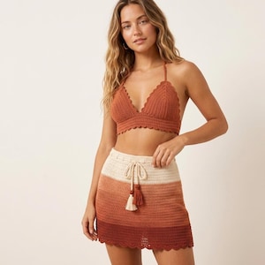May include: A rust-colored crochet bralette top with a scalloped edge and a matching crochet mini skirt featuring a horizontal ombre pattern in cream, rust, and dark brown, complete with a drawstring waist and two hanging tassels.