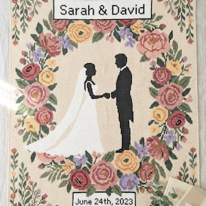 May include: A cream-colored woven blanket featuring a central floral wreath of pink, yellow, and purple flowers surrounding a silhouette of a couple holding hands. The text Sarah & David and June 24th, 2023 is embroidered in black rectangular frames.