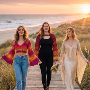May include: Three people walking on a wooden boardwalk near the beach at sunset. The person on the left wears a colorful crochet top and jeans. The middle person wears a red and black crochet top and black pants. The person on the right wears a cream-colored dress and crochet shawl.