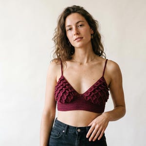May include: A burgundy crochet top with a V-neckline and ruffled detailing. The top has thin spaghetti straps and is paired with dark wash denim jeans. The top is a handmade garment.