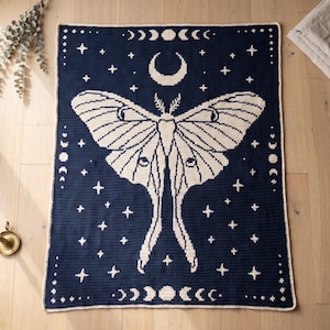May include: A rectangular crochet blanket featuring a large cream-coloured moth centred on a dark navy blue background. The design includes a crescent moon above the moth, stars scattered throughout, and a border of moon phases in cream yarn.