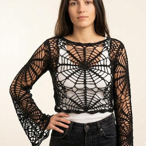May include: A black long-sleeved crochet top featuring an intricate spider web pattern throughout the garment. The piece has a round neckline, a cropped length, and flared sleeves, worn over a plain white tank top and dark denim jeans.